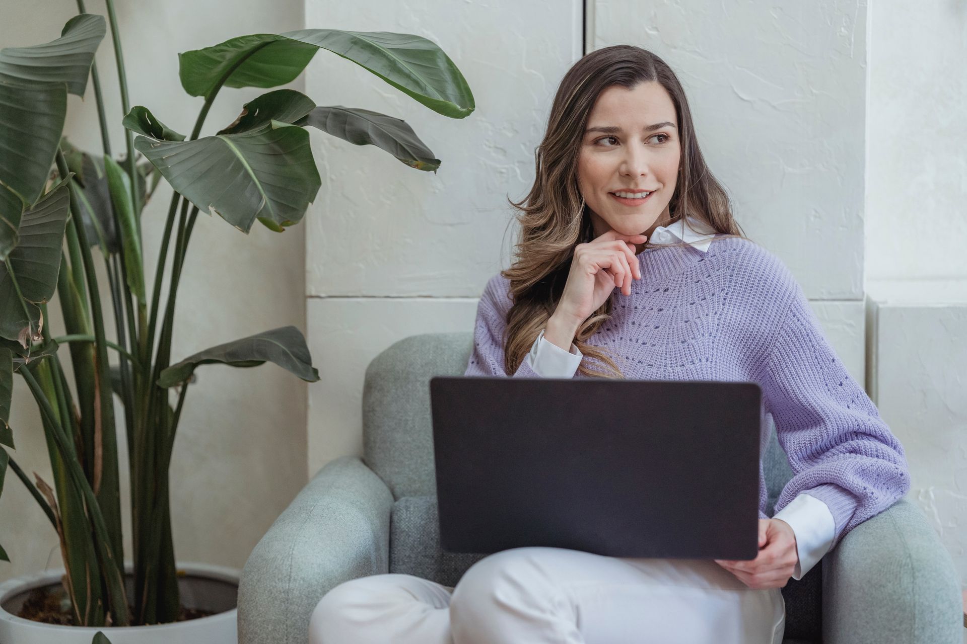 A woman sitting in a chair with a laptop, looking away thoughtfully as she thinks about how to find a job and plan her next career move.
