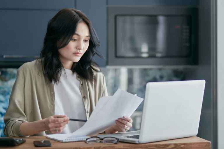 Professional woman reviewing printed resume documents while working at a wooden desk with a laptop.